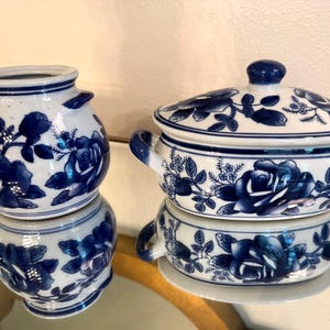 May include: Two pieces of white ceramic tableware with a blue floral design. One is a small jar with two handles, and the other is a lidded serving dish with handles. Both pieces have a glossy finish and are reflected on a mirrored surface.