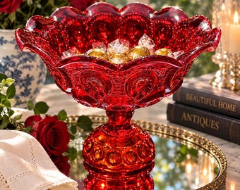 May include: A vibrant red glass pedestal bowl featuring a scalloped rim and a textured moon and star pattern. The bowl is filled with several gold-wrapped round candies and sits on a circular mirrored tray with a decorative gold metal edge.