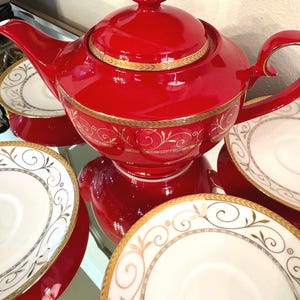 May include: A red teapot with gold trim and a matching set of saucers. The teapot and saucers feature a delicate gold floral design.