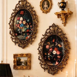 May include: A collection of ornate, gold-framed artwork and decorative pieces. Two large oval frames with floral paintings, a small portrait, a landscape painting, and a decorative shelf with a vase are displayed on a cream-colored wall.