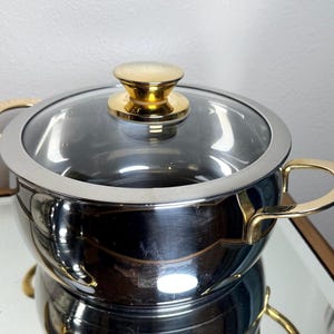 May include: Stainless steel cooking pot with a glass lid and gold-colored accents. The pot features a shiny, reflective surface, gold handles, and a gold knob on the lid. A modern kitchenware item.