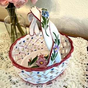 May include: A white ceramic basket with a lattice design and a red rim. The basket features a handle with floral accents in blue, green, and purple. Pink flowers in a glass vase are in the background.