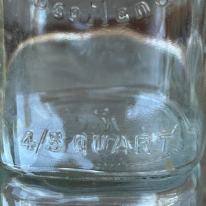 May include: Close-up of a clear glass bottle with the embossed word "Scotland" at the top. The bottle is marked "4/5 QUART" in the center. The glass has a slightly textured surface.