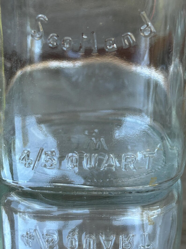 May include: Close-up of a clear glass bottle with the embossed word "Scotland" at the top. The bottle is marked "4/5 QUART" in the center. The glass has a slightly textured surface.