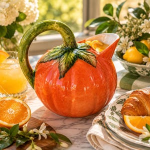 May include: A ceramic pitcher shaped like a bright orange pumpkin with a green stem handle and green leaves at the top. The pitcher has a rounded body, a wide pouring spout, and a glossy finish, filled with a yellow liquid and orange slices.