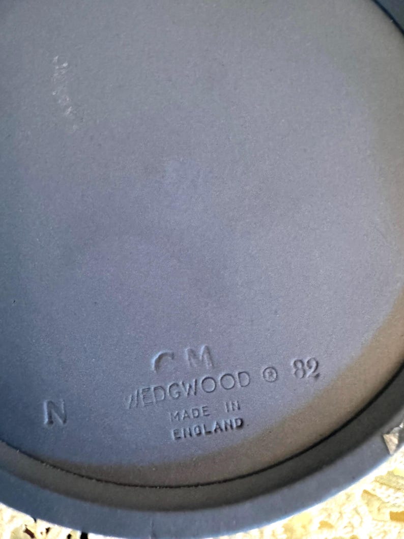 May include: Close-up of a blue Wedgwood ceramic piece. The text "CM WEDGWOOD &copy; 82 MADE IN ENGLAND" is stamped on the surface. The letter "N" is also visible. The piece has a matte finish.