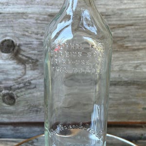 May include: Clear glass bottle with a weathered metal cap. The bottle has embossed text, including "John Walker & Sons Ltd" and "1/5 Quart". The bottle is rectangular with rounded shoulders.