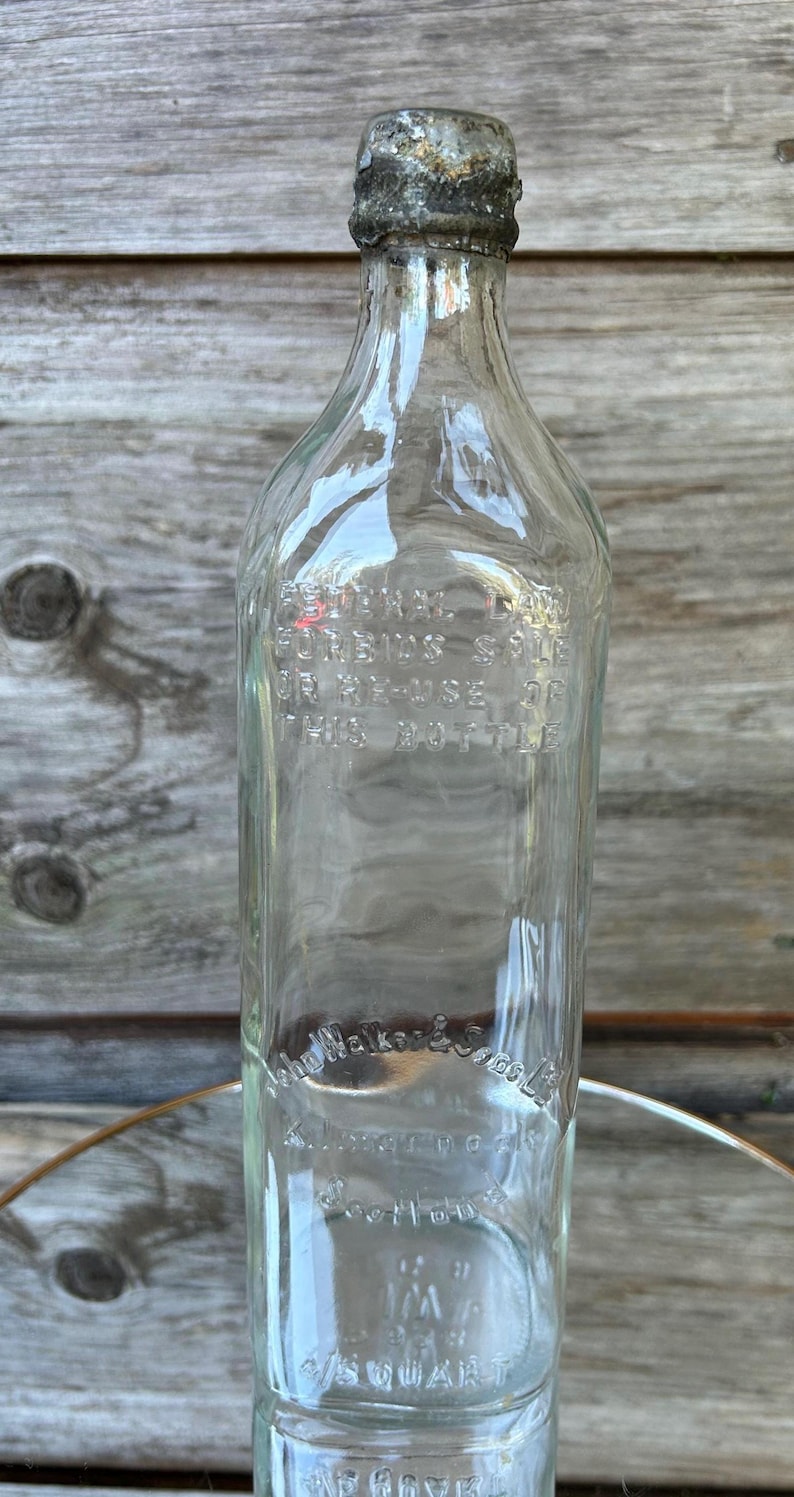 May include: Clear glass bottle with a weathered metal cap. The bottle has embossed text, including "John Walker & Sons Ltd" and "1/5 Quart". The bottle is rectangular with rounded shoulders.