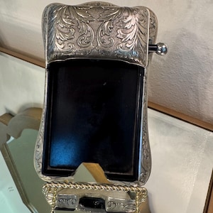 May include: A silver-toned, rectangular picture frame with ornate floral engravings. The frame has a black interior and a gold-colored rope-like detail at the base. A small, silver ball is attached to the side.