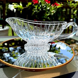 May include: A clear glass gravy boat with a handle and a spout, resting on a round mirror with a gold rim. The gravy boat has a textured, ribbed design. The background features greenery and red flowers.