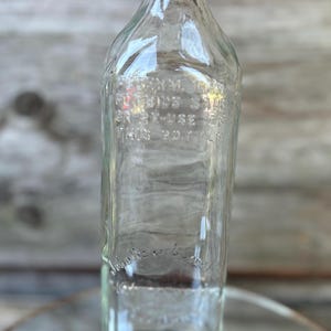 May include: Clear glass bottle with a metal cap. The bottle has embossed text that reads "Federal Law Forbids Sale or Re-Use of This Bottle." The bottle is rectangular with rounded shoulders.