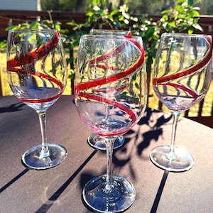 May include: Four clear wine glasses with a red swirl design. The glasses are on a dark surface, with sunlight and greenery in the background. The red design wraps around the glass.