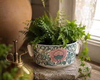 May include: A round ceramic bowl with two side handles, featuring a hand-painted pattern of a large pink flower surrounded by green swirling vines and leaves. The bowl contains a green fern and trailing ivy plant, sitting near a brass mister.