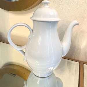 May include: A white ceramic teapot with a lid, curved handle, and spout. The teapot has a glossy finish and is reflected in the surface below. The design is simple and elegant, suitable for serving tea.