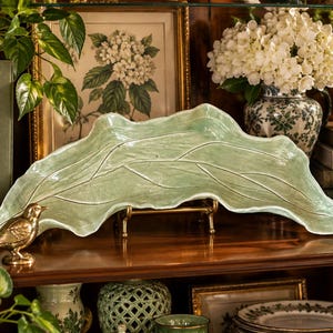 May include: A long, pale green ceramic serving tray shaped like a large, elongated leaf with prominent, raised vein details. The tray features a glossy, undulating surface with irregular, organic edges, displayed on a small gold metal stand.