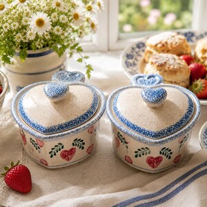 May include: Two heart-shaped ceramic lidded dishes with blue and white floral trim and heart designs. Strawberries, scones, and a vase of daisies are also visible, creating a charming, rustic aesthetic.