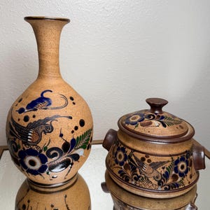 May include: A tan ceramic vase and a covered pot with hand-painted bird and floral designs in blue, black, and white. The vase has a slender neck and rounded body. The pot has handles and a lid with a knob.