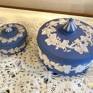 May include: Two blue ceramic lidded boxes with white relief grape vine designs. The boxes are different sizes, with a conical finial on each lid. The boxes are on a white lace tablecloth.