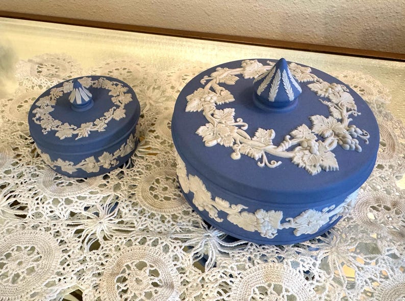 May include: Two blue ceramic lidded boxes with white relief grape vine designs. The boxes are different sizes, with a conical finial on each lid. The boxes are on a white lace tablecloth.