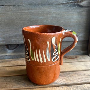 May include: A handmade, terracotta-colored mug with a rounded base and a handle. The mug is adorned with vertical painted designs in white, green, black, and gray. The mug is likely used for drinking beverages.