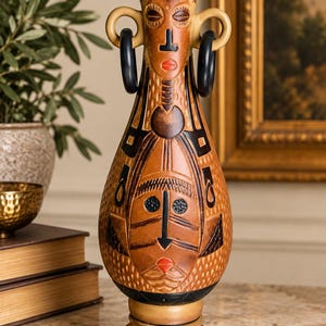 May include: A tall ceramic vase featuring a tan and brown finish with two stylized faces embossed on the front. The vase has two black hoop handles on the sides, a flared rim, and a black circular base decorated with small white dots and carvings.
