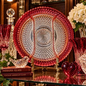 May include: A round, divided glass serving platter featuring a vibrant red outer rim with a diamond-cut pattern and a clear, sunburst-patterned center. The platter is supported by a gold-toned metal stand, showcasing intricate craftsmanship.