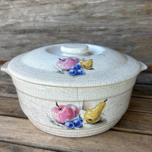 May include: Cream-colored ceramic casserole dish with lid. The dish features a fruit design with pink apples, yellow pears, and blue grapes. The lid has a small, round handle. The dish has a crackle glaze.