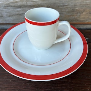 May include: A white ceramic mug with a red rim sits on a matching white plate with a red border. The mug has a curved handle. The plate and mug are on a dark wooden surface. This set is ideal for serving coffee or tea.