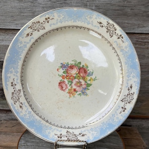 May include: A vintage porcelain plate with a light blue border featuring a floral and scroll design. The plate has a gold rim and a central floral bouquet in shades of pink, yellow, and blue. The plate is displayed on a metal stand.
