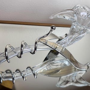 May include: A clear glass and silver wine bottle opener with a decorative floral design. The opener features a spiral corkscrew and a flower-shaped top. The opener is reflected in a mirrored surface.