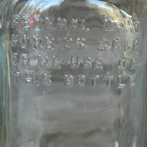 May include: Clear glass bottle with embossed text reading "FEDERAL LAW FORBIDS SALE OR RE-USE OF THIS BOTTLE." The bottle has a slightly curved shape and reflects the surrounding environment.