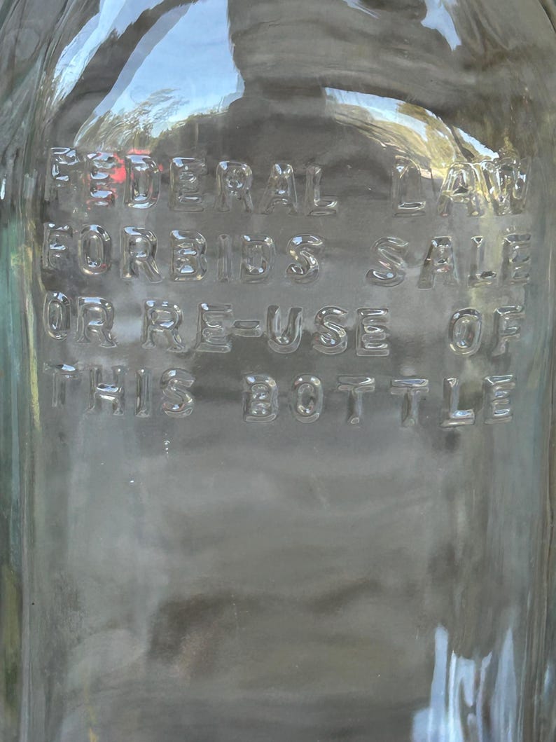 May include: Clear glass bottle with embossed text reading "FEDERAL LAW FORBIDS SALE OR RE-USE OF THIS BOTTLE." The bottle has a slightly curved shape and reflects the surrounding environment.