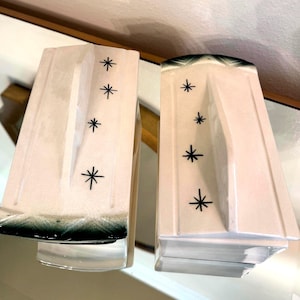 May include: Pair of vintage white ceramic bookends with a mid-century modern design. Each bookend features a starburst pattern in black and a dark green trim at the top. The bookends have a unique, architectural shape.