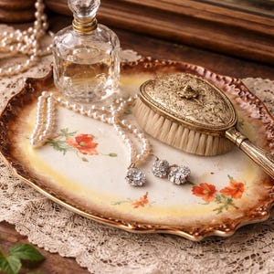 May include: An antique vanity tray with a floral design, holding a perfume bottle, a pearl necklace, earrings, and a decorative brush. The tray is approximately 12 inches long and 8 inches wide, with a gold-colored rim.