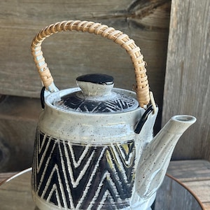May include: A ceramic teapot with a speckled gray finish and a black geometric pattern. The handle is made of woven light brown material. The lid is black with a small black knob. The teapot is sitting on a reflective surface.