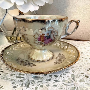 May include: A vintage teacup and saucer set. The cup and saucer are cream-colored with gold trim and a decorative scene of figures in period clothing. The saucer has a scalloped edge and floral accents. The teacup has a pedestal base and a gold handle.