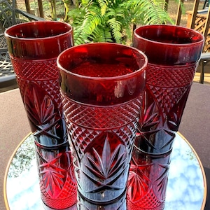 May include: Three ruby red drinking glasses with a cut glass design. The glasses are tall and cylindrical, with a diamond and leaf pattern. They are reflected in a round mirror.
