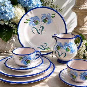 May include: A ceramic dinnerware set featuring a white base with hand-painted blue floral motifs and scalloped edges. The collection includes a large plate, a small pitcher, and two teacups with matching saucers, all accented with blue rims.