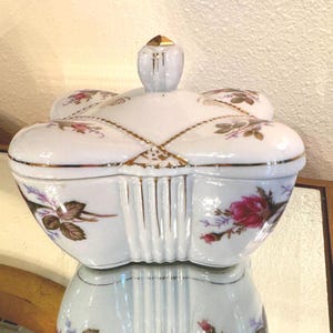 May include: A white porcelain trinket box with a floral design. The box has a unique, multi-lobed shape, with a matching lid and a small, gold-tipped finial. The box is decorated with pink roses and gold trim.