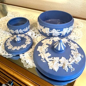 May include: A set of blue ceramic trinket boxes with white grape vine relief detailing. The set includes a small round box, a larger round box, and two lids. The lids have a central finial and grape vine relief.