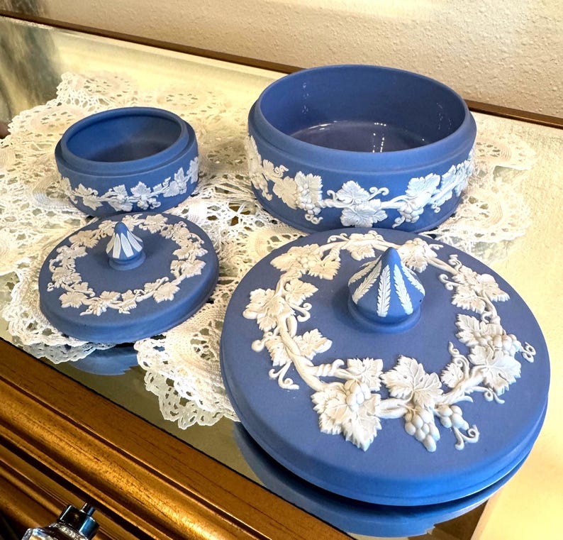 May include: A set of blue ceramic trinket boxes with white grape vine relief detailing. The set includes a small round box, a larger round box, and two lids. The lids have a central finial and grape vine relief.