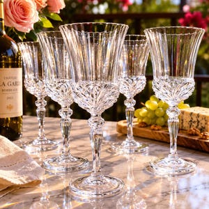 May include: A set of five clear crystal wine glasses featuring vertical fluted patterns on the bowls and stems, with intricate pressed geometric detailing around the base of each bowl, arranged on a white marble surface with a wine bottle.