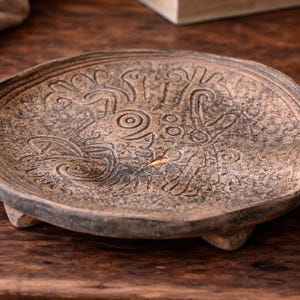 May include: A handcrafted, brown ceramic plate with an embossed design. The plate has a raised rim and rests on three small feet. The surface features an intricate pattern of stylized figures and shapes, perfect for decorative use.