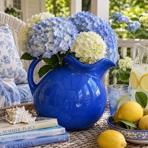 May include: A glossy cobalt blue ceramic pitcher filled with a large bouquet of white and light blue hydrangea flowers. The pitcher sits next to a stack of three books, a white seashell, a glass of water with lemon, and a bowl of fresh lemons.