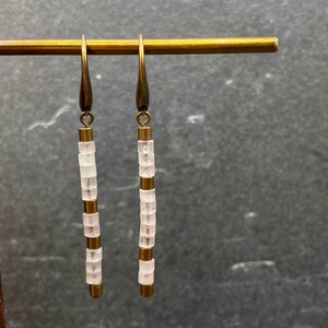 May include: A pair of long, gold-toned earrings with white, square-shaped beads. The earrings are hanging from a gold-toned bar.
