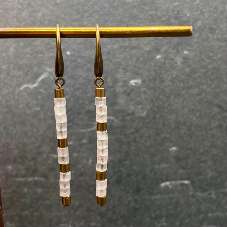 May include: A pair of long, gold-toned earrings with white, square-shaped beads. The earrings are hanging from a gold-toned bar.