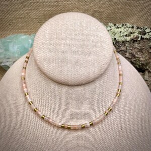May include: A delicate necklace made with small, light pink and white gemstone beads interspersed with gold beads. The necklace is displayed on a fabric-covered mannequin.