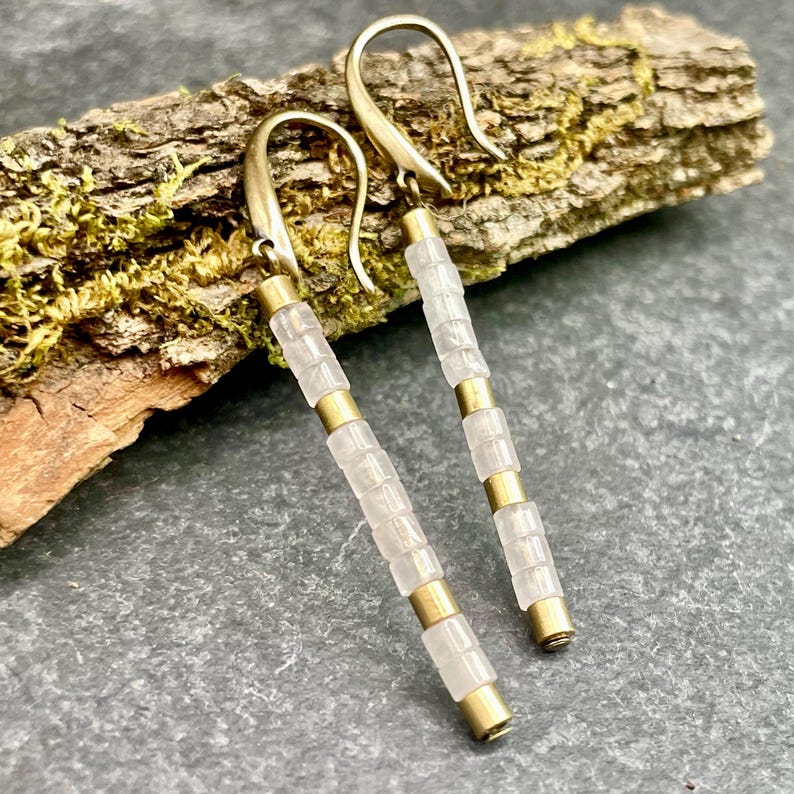 May include: A pair of long, gold earrings with white beads. The earrings are hanging from a wooden surface.