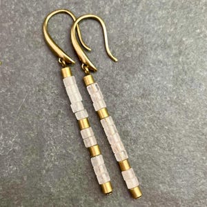 May include: A pair of long, gold dangle earrings with clear, faceted, square-shaped beads. The earrings have a simple, minimalist design.