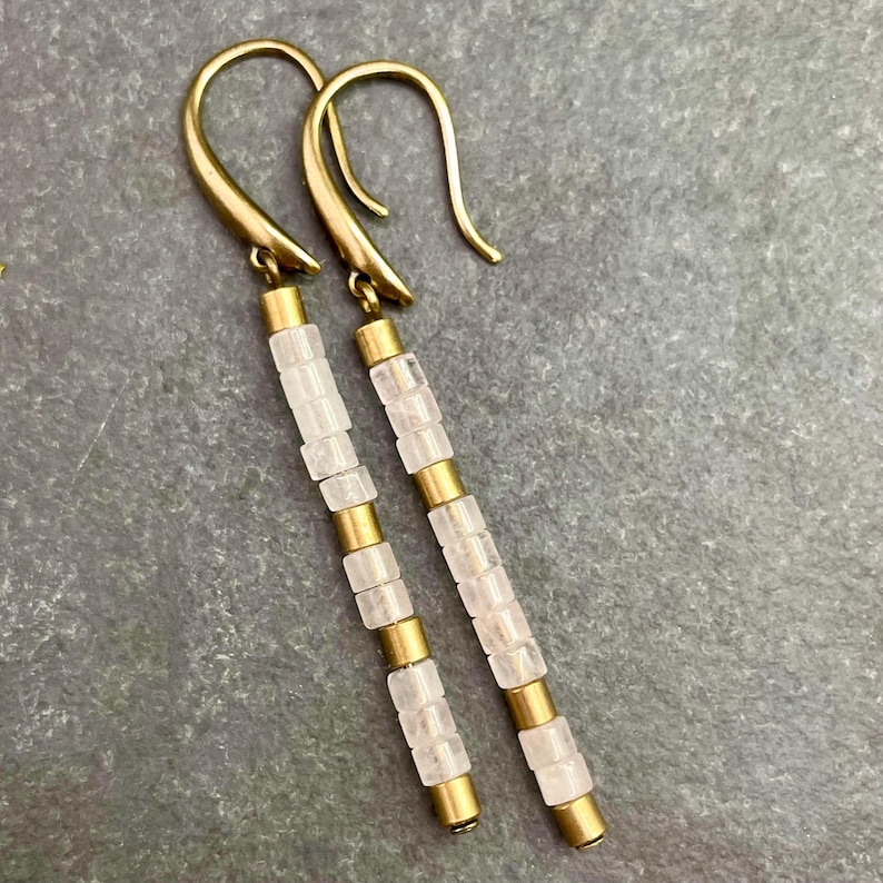 May include: A pair of long, gold dangle earrings with clear, faceted, square-shaped beads. The earrings have a simple, minimalist design.